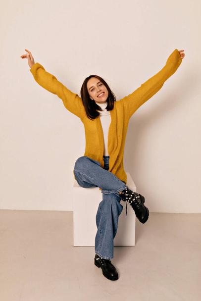 Full-lenght photo of adorable energy girl with dark hair wearing yellow shirt and jeans sitting on chair with raised up hands on isolated background.  - Foto, Bild