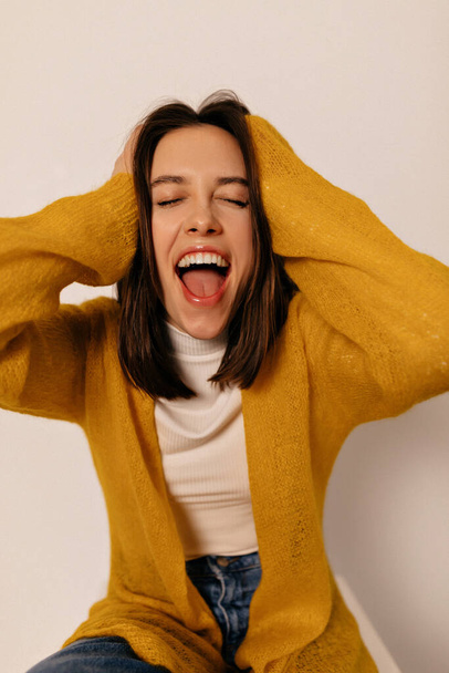 Excited lovely girl with dark hair is touching her head and screaming with teeth over isolated background. She wears yellow shirt  - Foto, imagen