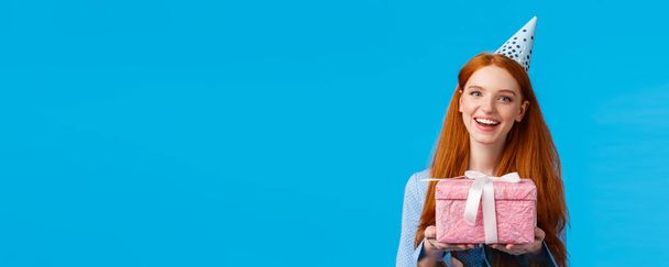 Tijd voor nu. Vrolijke roodharige tiener, college meisje vieren haar verjaardag, met schattige roze verpakt cadeau en het dragen van b-day hoed, vrolijk glimlachen staande blauwe achtergrond. - Foto, afbeelding