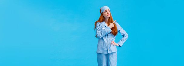 Uneasy and worried, concerned upset redhead female in nightwear and sleep mask, looking pointing upper left corner sad and distressed facing hard choice, standing troubled blue background. - Photo, Image