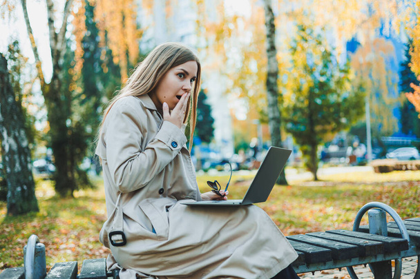 Urocza, jasnowłosa młoda kobieta siedzi w jesiennym parku na ławce i pracuje nad laptopem. Praca zdalna - Zdjęcie, obraz