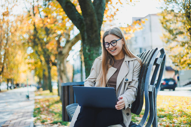 Een schattige blonde jonge vrouw zit in een herfstpark op een bankje en werkt aan een laptop. Afstandswerk - Foto, afbeelding