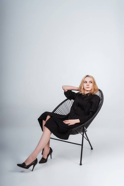 girl in a black dress on a white background. girl sitting on a knitted black chair on a white cyclorama. photoshoot of black clothes with a model - Photo, Image