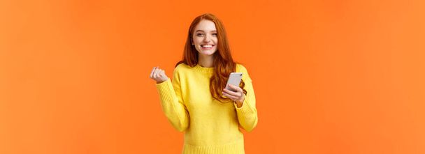 Happy girl share good news with you. Cheerful pretty redhead female holding smartphone, fist pump in triumph or celebration, winning in game, got message from guy she likes, orange background. - Photo, Image