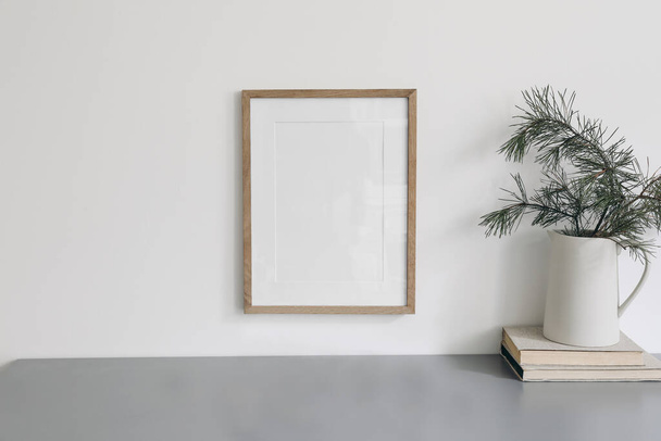 Christmas interior decor. Blank wooden picture frame mockup, white wall. Green pine tree branches in jug and old books. Grey working table. Old books. Winter home office still life, poster display. - 写真・画像