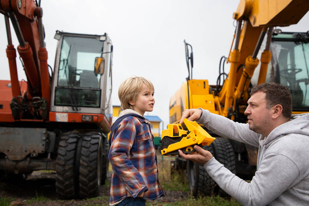 Papá e hijo, en el contexto de los equipos de construcción excavadoras, están examinando con entusiasmo una excavadora de juguete, que un hombre tiene en sus manos. El interés del niño en las máquinas y su trabajo - Foto, imagen