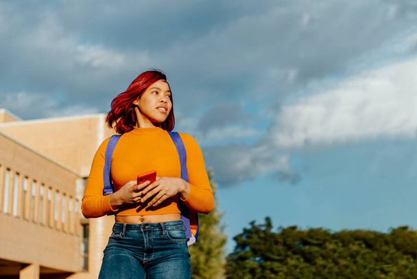 jong latina meisje met rood haar, rugzak, lopend met haar smartphone in haar handen aan de rand van de universiteitscampus. student die de klas verlaat. - Foto, afbeelding