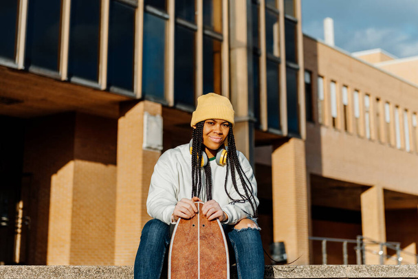 portrait of latina girl with braids and vitiligo, with wool cap and headphones sitting holding a longboard skateboard outside high school. youth culture - Photo, Image