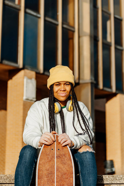 portrait looking at camera of young and latin girl with vitiligo, braids and yellow wool cap, sitting and holding a skateboard in her hands. - Photo, Image