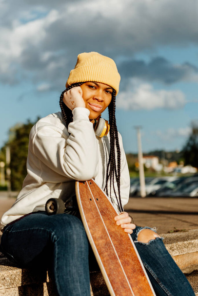 portrait of Hispanic girl with vitiligo, sitting holding a longboard skateboard. Latina girl with braids looking at camera. youth culture. - 写真・画像