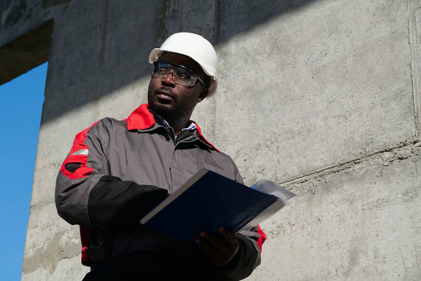 Αφροαμερικάνος εργάτης στέκεται στο εργοτάξιο με τα έγγραφα εργασίας και κοιτάζει γύρω - Φωτογραφία, εικόνα