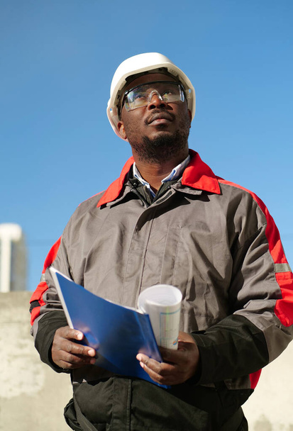 Un travailleur afro-américain se tient sur le chantier, tient dans les mains des documents de travail et regarde autour de lui - Photo, image