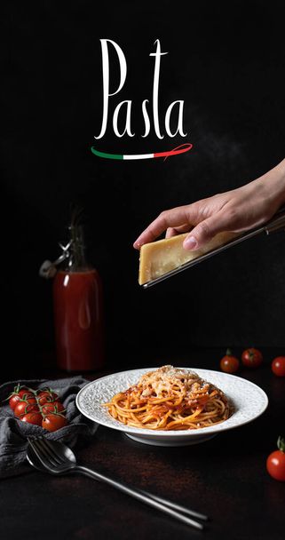 Nudeln mit Tomatensauce und Parmesan. Weißer Teller mit Spaghetti auf dunklem Hintergrund mit dem Wort Pasta. Hände reiben Parmesan auf Spaghetti. Traditionelle italienische Küche  - Foto, Bild