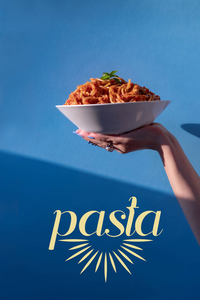 Eine weibliche Hand hält einen Teller Spaghetti mit Tomatensauce auf einem weißen Teller vor blauem Hintergrund mit dem Wort Pasta. Das Konzept der modernen italienischen Küche. Selektive Fokussierung aus nächster Nähe - Foto, Bild