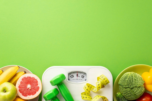 Detox concept. Top view photo of plates with fruits and vegetables grapefruit apple bananas pepper cabbage cucumber dumbbells and scales on isolated green background with empty space - Photo, Image