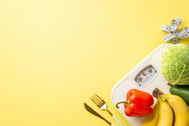 Diet concept. Top view photo of vegetables and fruits cabbage avocado bell pepper bananas cutlery tape measure and scales on isolated pastel yellow background with empty space - Photo, Image