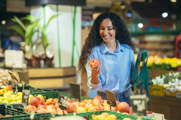 A beautiful young Hispanic woman is shopping in a supermarket. She stands smiling at the vegetable and fruit section, picks and holds an apple in her hands, puts it in an eco-bag. - Valokuva, kuva