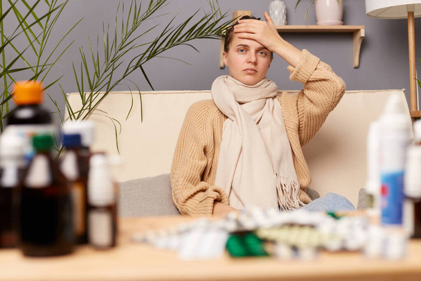 Image of sick unhealthy woman sitting on sofa wrapped in scarf surrounded with medicines, feels unwell, having headache and high temperature, touching her forehead, posing in living room at home. - Foto, imagen