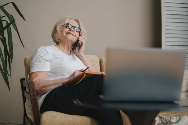 Ευτυχισμένη ώριμη ηλικιωμένη γυναίκα που χρησιμοποιεί συσκευές laptop και τηλεφώνου ξοδεύοντας ελεύθερο χρόνο με την τεχνολογία στον καναπέ στο σύγχρονο σαλόνι διαμέρισμα. Ηλικιωμένοι online γενιά - Φωτογραφία, εικόνα