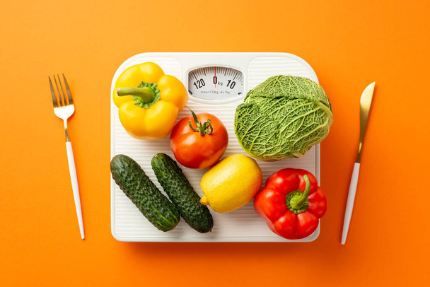 Proper diet concept. Top view photo of vegetables on scales knife and fork on isolated orange background - Фото, зображення