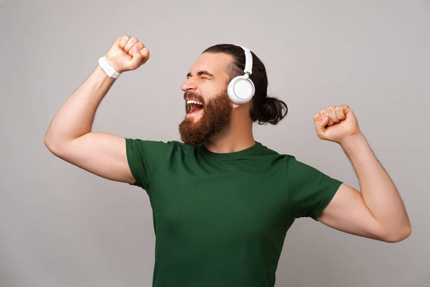 Knappe man draagt groen t-shirt geniet van muziek via draadloze koptelefoon over grijze achtergrond in een studio. - Foto, afbeelding