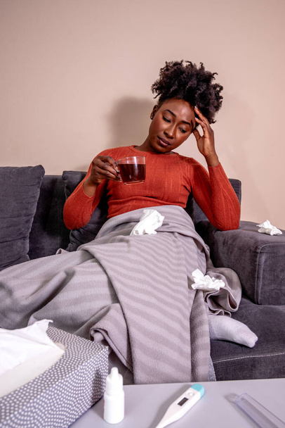 Kobieta siedząca na kanapie z filiżanką herbaty, źle się czuje. Afroamerykanka ma migrenę, trzyma się za czoło. - Zdjęcie, obraz