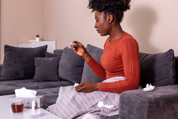 Woman sitting on a sofa, feeling sick. African American girl having flu, and checking her body temperature. - Foto, Bild