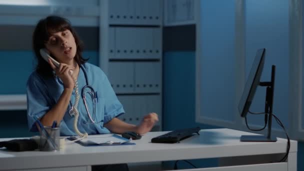 Lékař sestra zvedá pevnou linku telefon diskutovat hlášení pacientů s dálkovým lékařem plánování léčby k vyléčení onemocnění. Lékařská asistentka pracující přes hodiny v nemocniční kanceláři - Záběry, video
