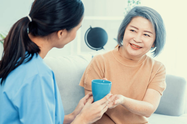 Γυναίκα φροντίστρια που σερβίρει τον ικανοποιημένο ασθενή της με ένα φλιτζάνι καφέ στο σπίτι, χαμογελώντας ο ένας στον άλλο. Ιατρική περίθαλψη για συνταξιούχους, Υπηρεσία κατ 'οίκον υγειονομικής περίθαλψης. - Φωτογραφία, εικόνα