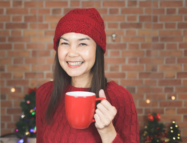 Kırmızı örgü süveter ve şapkalı Asyalı bir kadının portresi mutfakta duruyor Noel ağacıyla süslenmiş kırmızı bardaktan kahve içiyor, mutlu bir şekilde gülümsüyor ve kameraya bakıyor. Kış ve Noel. - Fotoğraf, Görsel