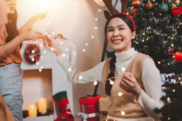 Group of happy Asian friends celebrating Christmas and decorate the Christmas tree indoors. Beauty woman with Christmas Gifts. New Year party. Woman hands decorate Christmas tree red ball, bauble. - 写真・画像