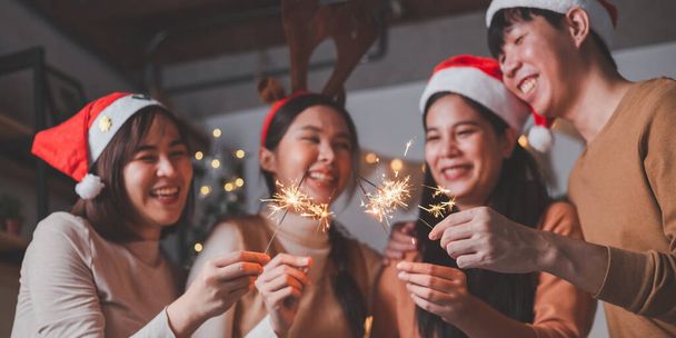 Eine Gruppe junger asiatischer Männer und Frauen als Freundinnen, die sich bei einer Neujahrsfeier amüsieren und Wunderkerzen bei einer mitternächtlichen Countdown-Party zu Hause mit Christbaumschmuck für die Weihnachtszeit hochhalten. - Foto, Bild
