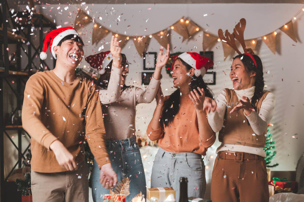 Bir grup yetişkin Asyalı genç arkadaş Noel arifesinde eğleniyor ve evde Noel ağacı ve süslemelerle konfeti üflüyorlar. Yeni yıl, tatil, kutlama partisi Arkadaşlar eğleniyor. - Fotoğraf, Görsel