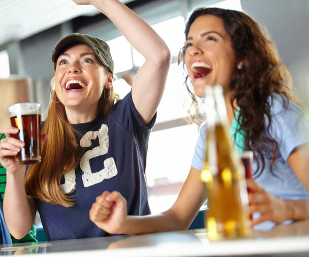 Wat zou ze doen zonder drank en sport. Twee vriendinnen juichen hun favoriete team toe terwijl ze achterover leunen met bier aan de bar. - Foto, afbeelding