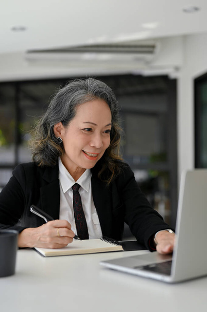 Professional Asian aged businesswoman working at her desk, using laptop computer and taking notes on her notepad. - 写真・画像