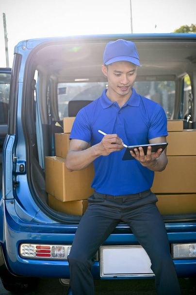 Toimittaja sinisessä univormussa tarkistaa paketin tabletin ennen lähettämistä asiakkaalle, istuu hänen pakettiautonsa  - Valokuva, kuva
