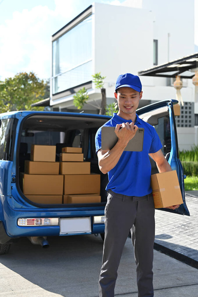 Young Asian delivery man checking orders with tablet before handing package to customers while standing in front of his van  - Photo, Image