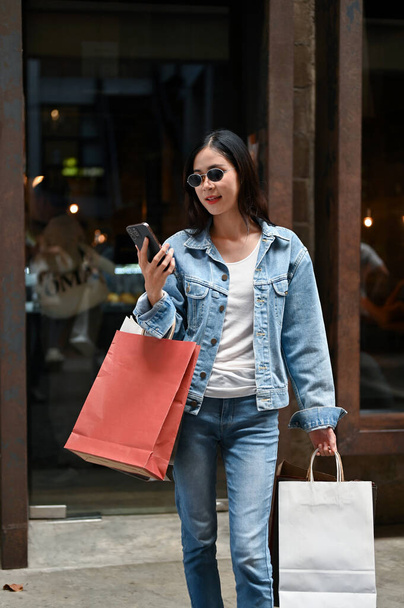 Porträt: Junge Asiatin hält Smartphone in der Hand und trägt Einkaufstüten, während sie im Stadtzentrum einkauft. - Foto, Bild
