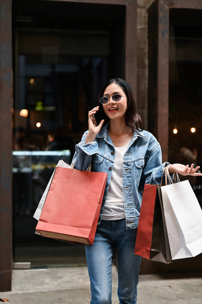 ショッピングバッグを持っているエレガントで美しいアジアの若い女性は、商店街を歩いている間に電話で話すのを楽しんでいます. - 写真・画像