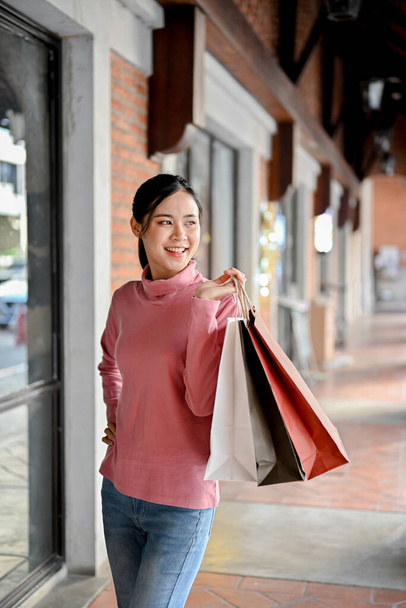 Portret, ładna i czarująca młoda Azjatka niosąca torby na zakupy, stojąca przed wejściem do sklepu. - Zdjęcie, obraz