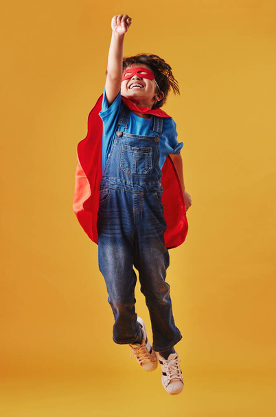 Adorable little asian boy wearing a mask and a cape while pretending to be a superhero against an orange studio background. Cute happy boy pretending to be a character for halloween. - Photo, image
