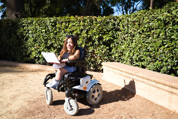 jonge gehandicapte vrouw in rolstoel en beperkte mobiliteit met haar laptop computer telewerken in het park. - Foto, afbeelding