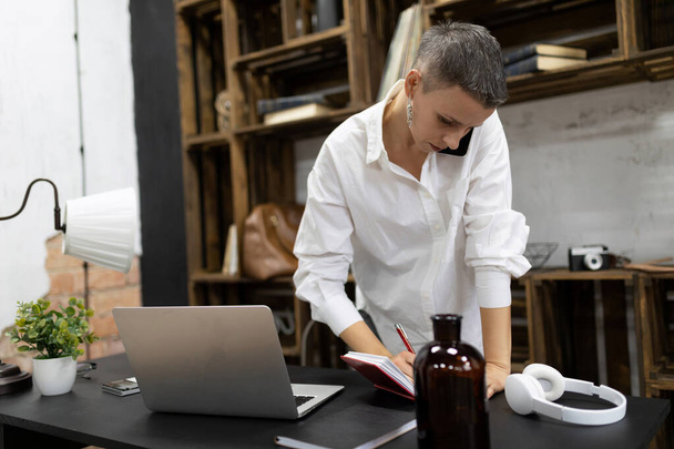 kobieta z krótką fryzurą pracuje w biurze rozmawiając przez telefon robiąc notatki w notatniku. - Zdjęcie, obraz