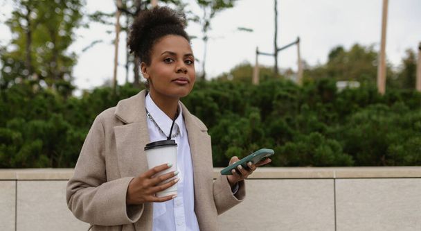 élégante femme d'affaires afro-américaine avec une tasse de café et un téléphone dans ses mains à l'extérieur. - Photo, image