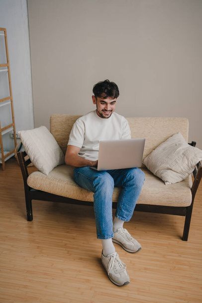 Młody człowiek pracujący na laptopie, relaksujący się na wygodnej sofie w domu. Komunikacja internetowa. Szczęśliwy styl życia. - Zdjęcie, obraz