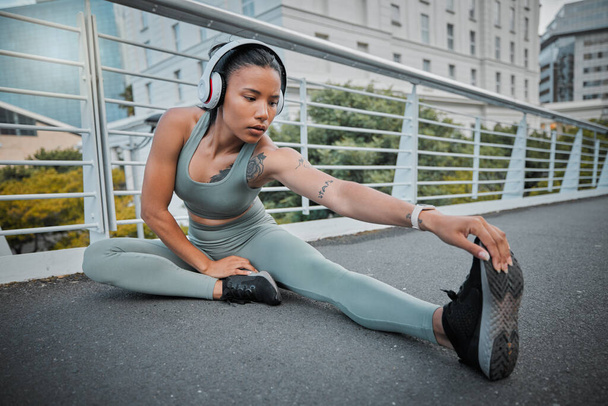 Young mixed race female athlete wearing headphones and listening to music while stretching before a run outside In the city. Confident hispanic sportswoman warming up before starting her exercise. - Photo, Image
