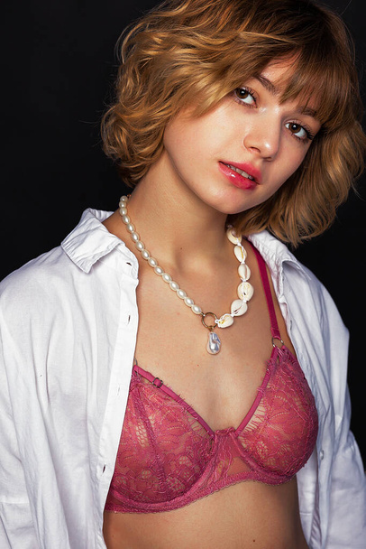 Caucasian woman with slender body posing in studio in white shirt and pink bra with a necklace, a white jewel around her neck. Isolated over black background. - Фото, изображение