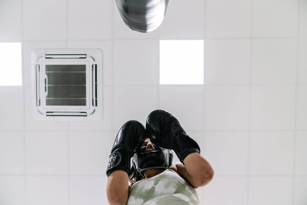 Vista de baixo de uma jovem mulher usando luvas de boxe pretas na posição de guarda na frente de um saco de perfuração em um ginásio - Foto, Imagem