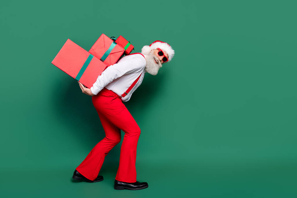 Full length photo of cool positive man dressed xmas costume holding back heavy gifts stack empty space isolated green color background. - Foto, afbeelding