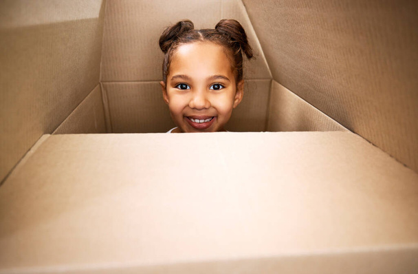 Portrait of a cute little hispanic girl playing with a cardboard box in a new apartment. Cute mixed race girl hiding in a box and smiling in a house. Relocation and moving day of big family. - Fotó, kép